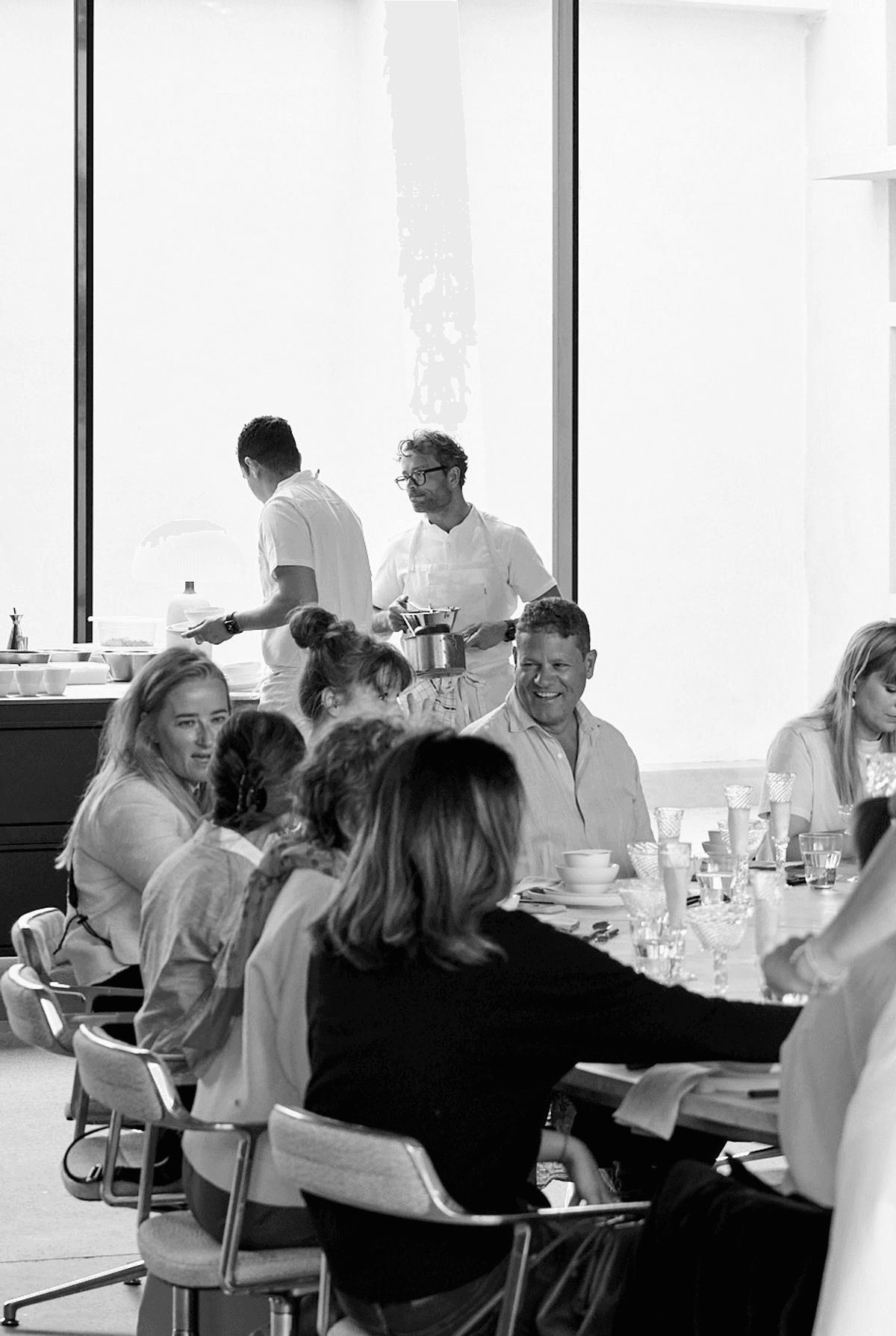 Black-and-white photo of guests at a table during an event at Mogens Dahl Concert Hall, with chefs in the background preparing and serving a carefully selected menu. The focus is on creating a seamless experience with top-quality ingredients in an exclusive setting.