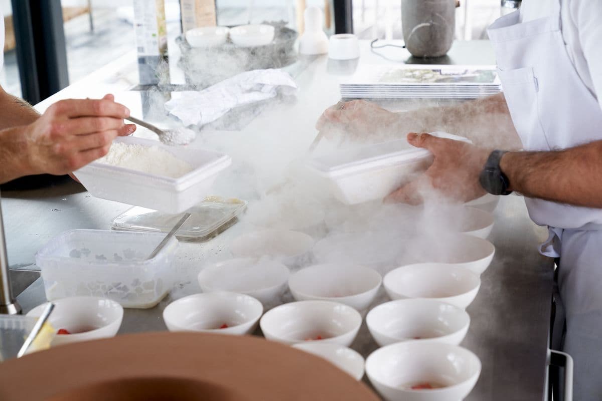 Close-up of hands from two chefs plating desserts on white deep plates in the kitchen at Mogens Dahl Concert Hall. White smoke from the cold creates a dramatic effect around the plates.