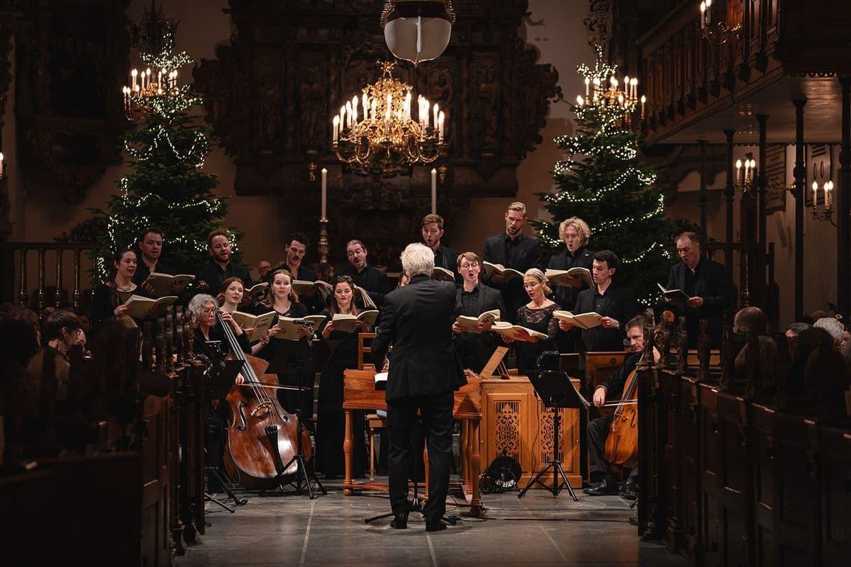 Mogens Dahl Kammerkor optræder til en koncert med julepynt i baggrunden. Dirigenten Mogens Dahl ses med ryggen til, mens han leder koret med præcision. Billedet illustrerer den festlige stemning og musikalske kvalitet, som har været fremhævet i anmeldelser af både Mogens Dahl Koncertsal og Kammerkor.