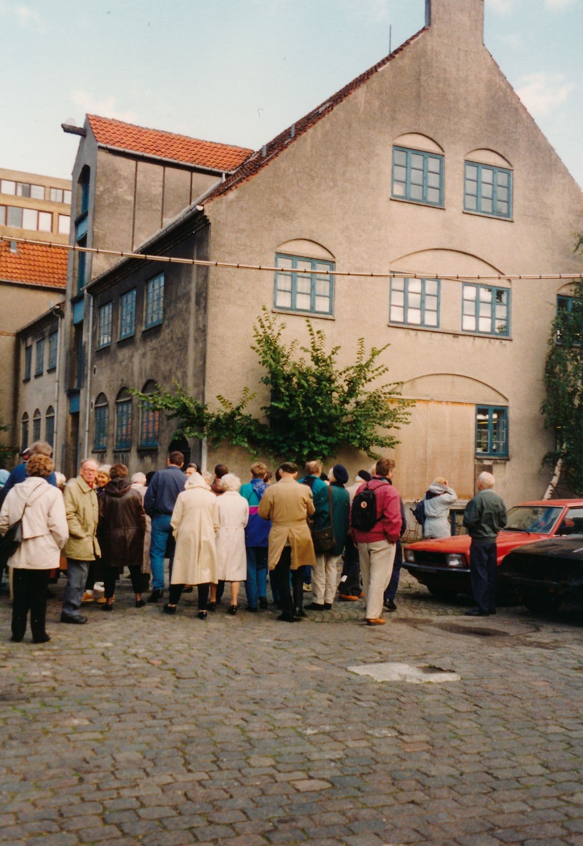 Historical photo of the courtyard at Mogens Dahl Concert Hall with people and a parked car from an earlier time. The image shows the old courtyard at Islands Brygge before it was transformed into the concert hall, providing insight into the site's previous function as a garage and workshop.