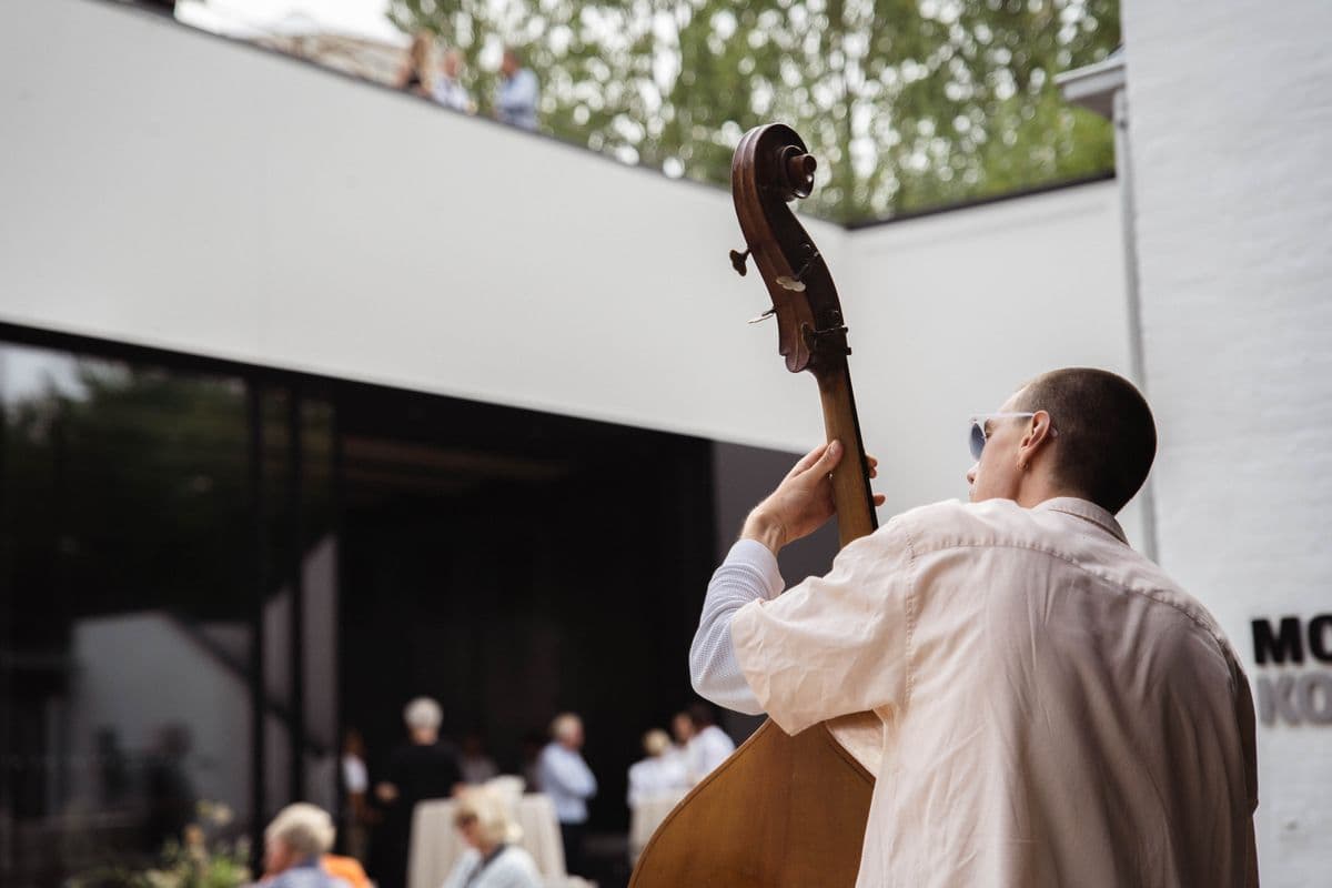 Photo of a person playing the cello outdoors at an event at Mogens Dahl Concert Hall. The image captures the musician in action while performing for guests in a beautiful outdoor setting.